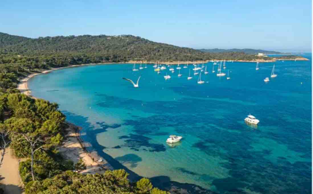 A coastal view features a calm bay surrounded by lush greenery and hills. Several boats are anchored in the clear blue water, creating a serene atmosphere. Soft sands line the shore, inviting leisurely activities by the beach, with distant hills providing a natural backdrop.