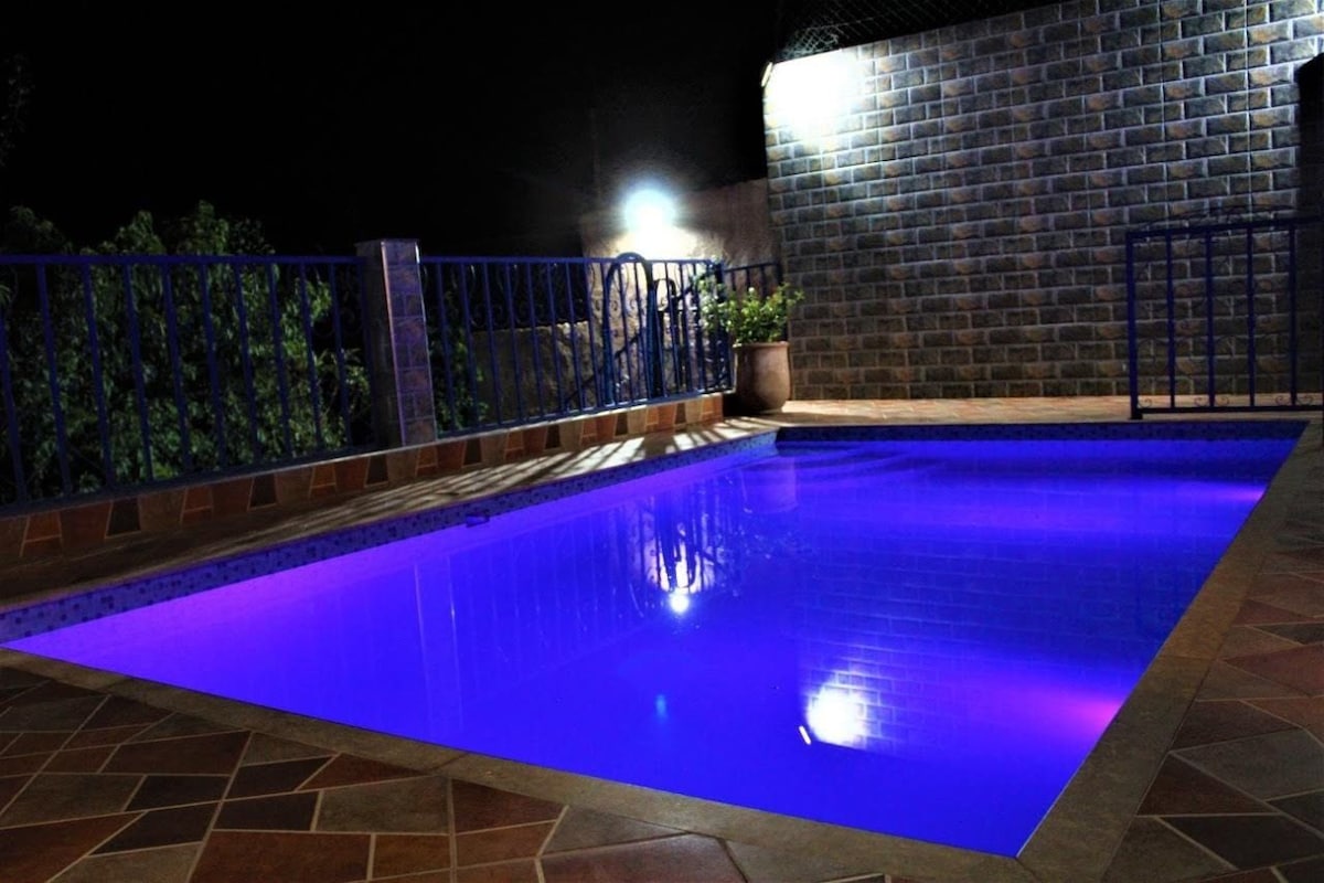 A calm swimming pool is illuminated in soft purple lighting at night, surrounded by a tiled patio. The pool is framed by a stone wall and decorative plants, creating a serene ambiance.