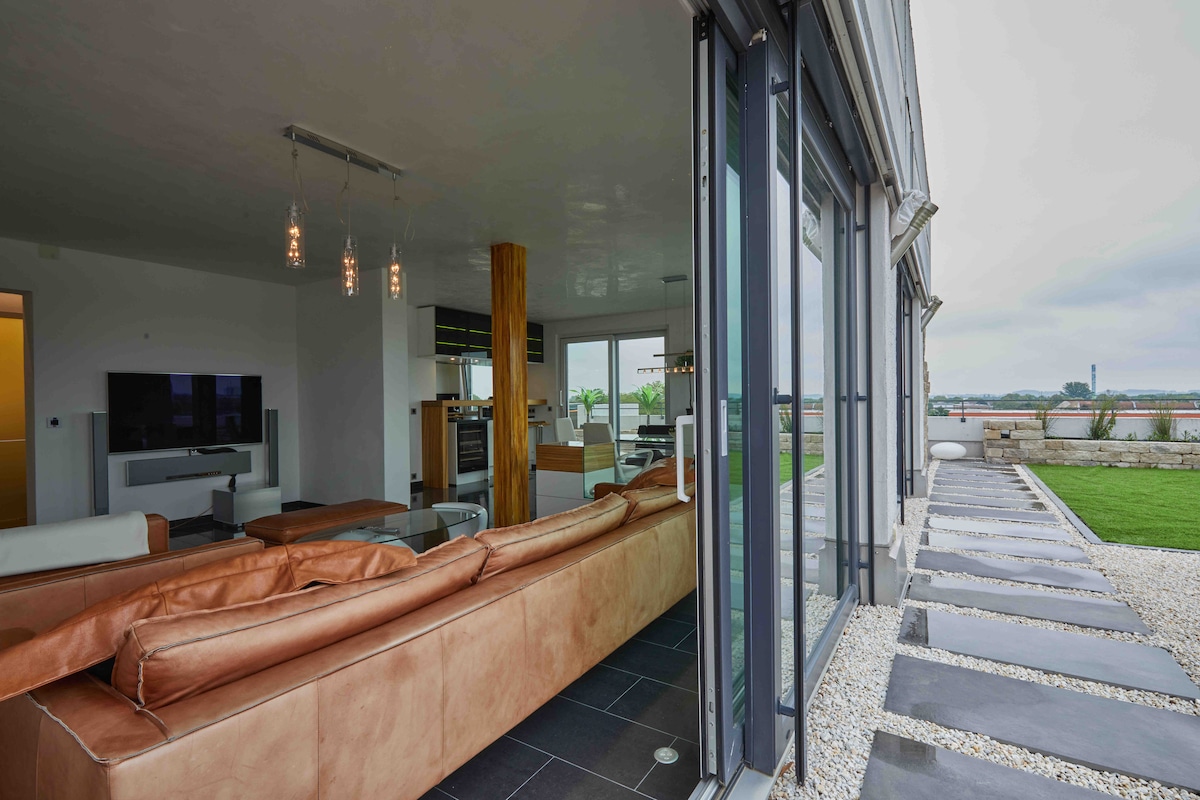 A spacious living area features a large leather sectional sofa, set against expansive glass doors that connect to an outdoor space. Sunlight fills the room, highlighting the modern decor and creating a seamless transition to the terrace, with views of the landscape beyond.