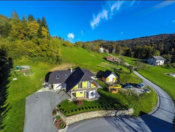 Aerial view of a charming property surrounded by lush greenery and hills. The main building features a yellow exterior with a dark roof, accompanied by several smaller structures and vehicles. A circular driveway is visible, along with outdoor spaces for relaxation.