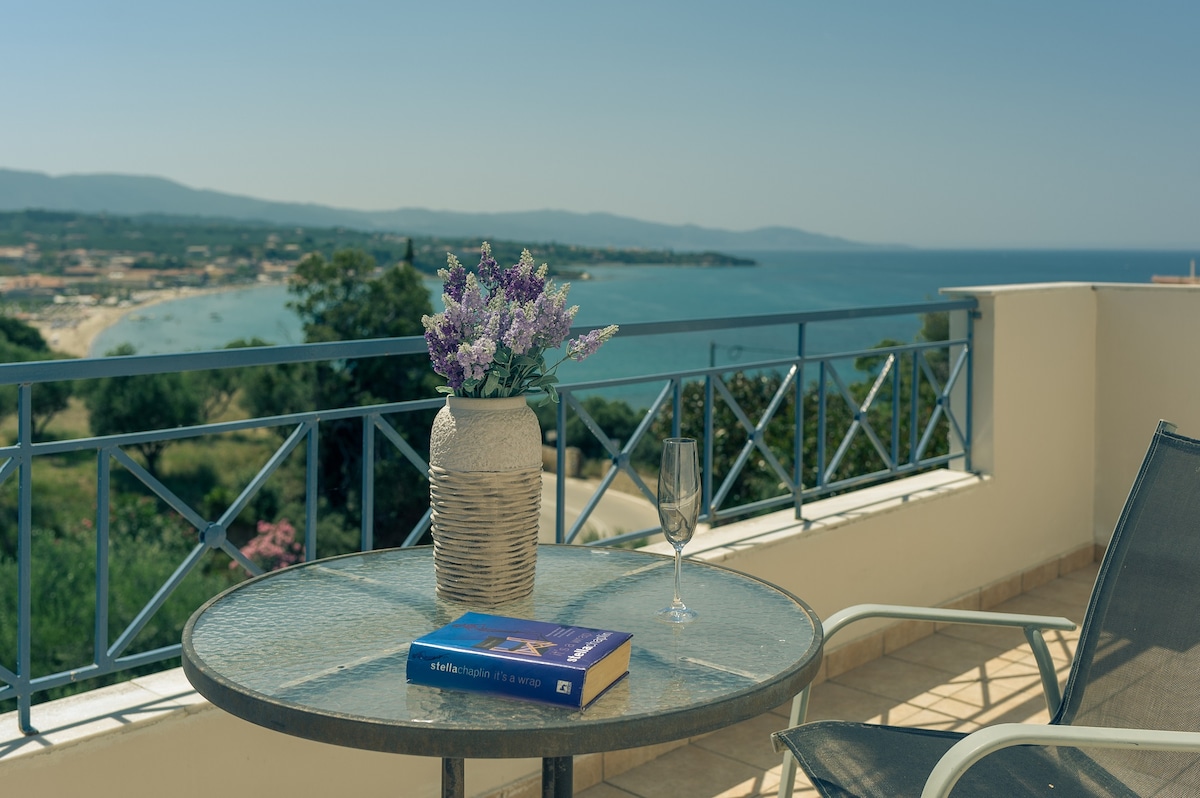 A balcony view showcases a glass table with a woven vase filled with lavender. A book rests on the table beside a sparkling wine glass, with panoramic views of the coastline and distant mountains visible in the background.