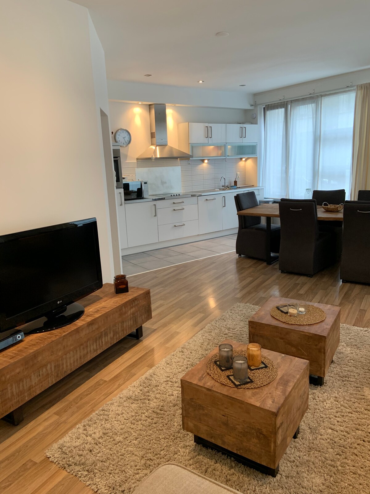 A modern living area is displayed, featuring a television on a wooden stand, accompanied by two square coffee tables. A dining table with dark chairs is visible in the background, with a well-equipped kitchen that includes built-in appliances and ample storage. Natural light fills the space through large windows covered by sheer curtains.