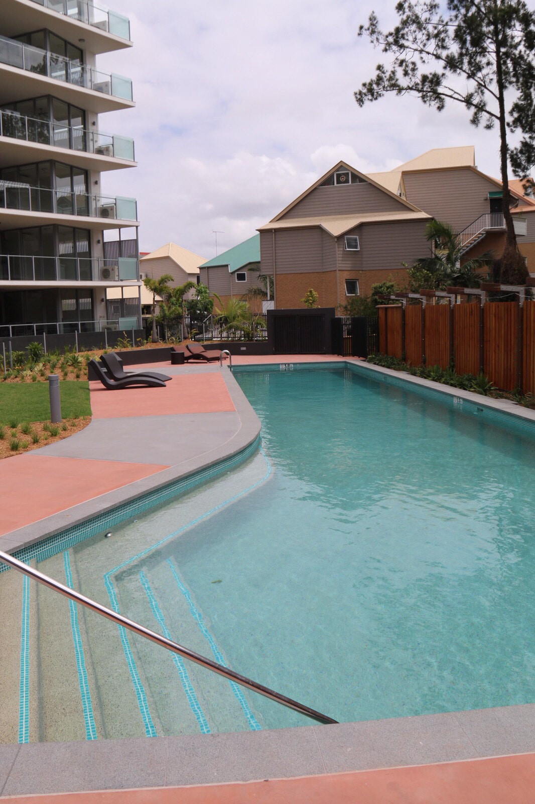 A large swimming pool is surrounded by well-maintained landscaping and wooden fencing. Lounge chairs are positioned nearby for relaxation. The pool features a gentle slope at one end leading into the water, reflecting the sky and nearby structures.