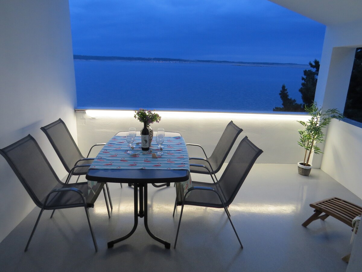 A spacious dining area is set with a large table and six chairs, positioned on a terrace overlooking the sea. Soft lighting highlights the serene space, complemented by a potted plant. The calm water reflects the evening sky's hues.