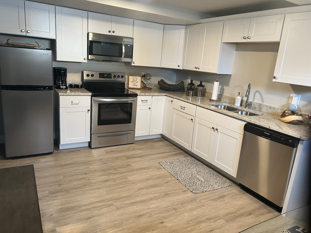 A modern kitchen is presented, featuring stainless steel appliances, including a refrigerator, microwave, and oven. White cabinetry and a light countertop provide a clean look. A decorative rug is placed in front of the sink, where natural light is filtered through a nearby window.