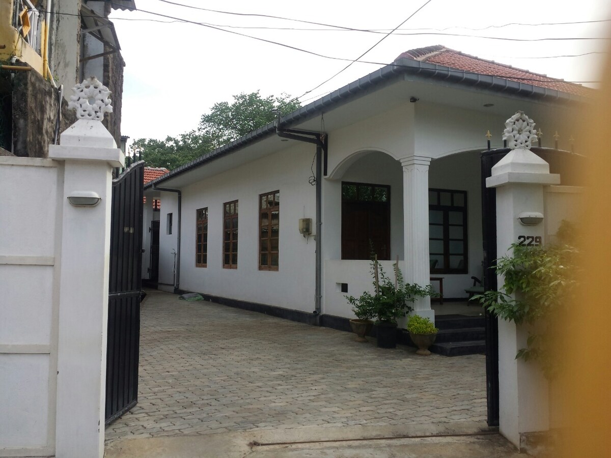 The exterior of a two-story villa is shown, featuring a clean, white facade and a terracotta roof. A paved driveway leads to the entrance, flanked by decorative potted plants. Large windows provide a glimpse of the interior, while the gated entrance offers privacy and security.