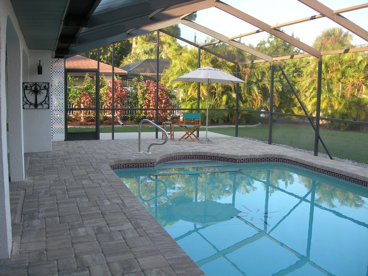 A serene outdoor pool area is enclosed by a screened lanai, featuring a clear blue pool reflecting the surroundings. A comfortable chair sits beside the pool, complemented by a shaded umbrella. Lush greenery and colorful plants create a calming backdrop.