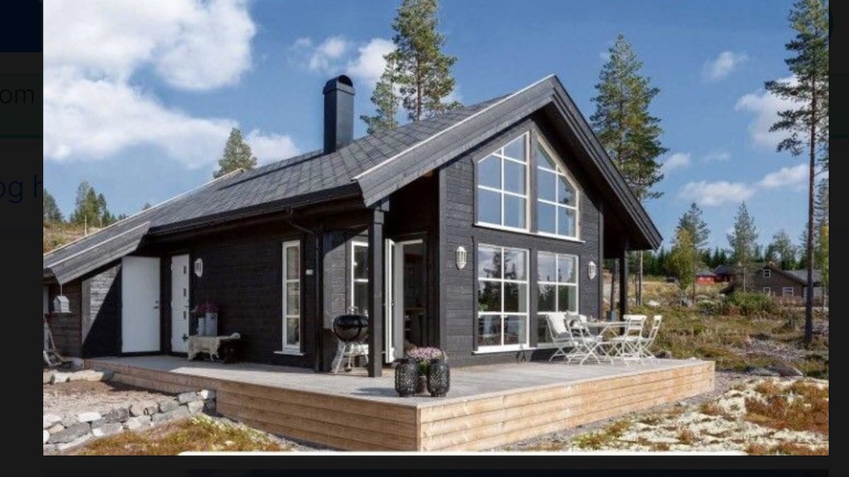 The cabin features a contemporary design with large windows that allow natural light to fill the interior. A spacious wooden terrace is visible, complete with a dining table and chairs. Surrounding greenery and hillside create a serene outdoor setting.