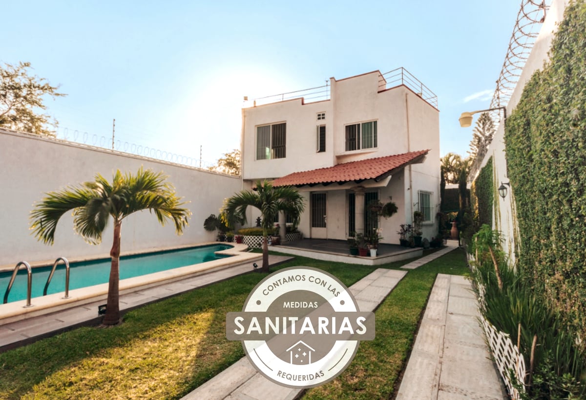 The exterior of the house showcases a clean white structure with a red-tiled roof, surrounded by green landscaping. A pool is visible to the left, with stepping stones leading to the entrance. Natural light brightens the scene, highlighting the inviting atmosphere.