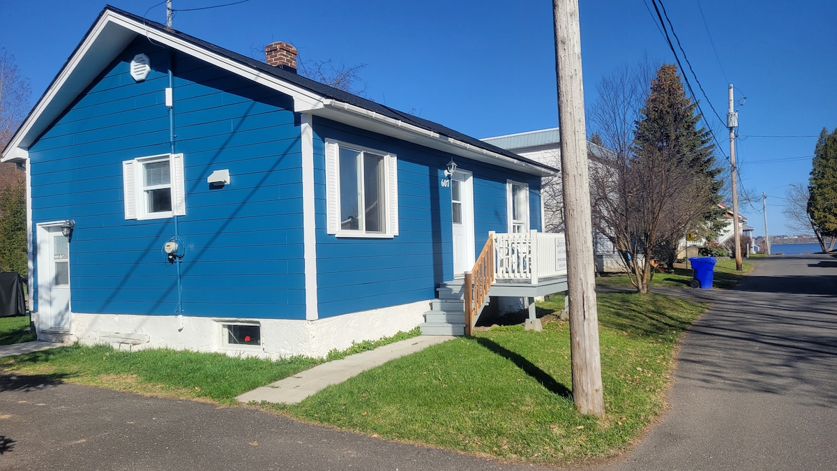 A charming blue house is situated on a well-maintained lot. The structure features multiple windows and a welcoming front porch with steps leading to the entrance. Lush green grass surrounds the home, with utility poles and trees providing a natural setting.