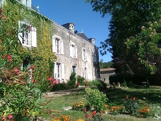 A historic house is framed by lush greenery and vibrant flowers, with large windows reflecting the sunlight. The exterior features stone walls partially covered in climbing plants, while the surrounding garden is adorned with colorful blooms and shaded by nearby trees.
