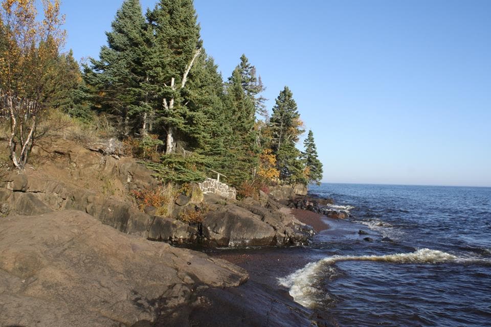 The image depicts a rocky shoreline adjacent to calm waters. Tall green trees line the edge, with autumn foliage visible. The soothing waves gently lap against the shore, while a clear blue sky provides a serene backdrop.