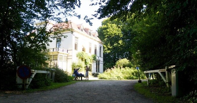 A historic country house is framed by lush greenery and large trees. A gravel path leads to the entrance, which features a covered terrace adorned with climbing plants. Light filters through the leaves, creating a serene and inviting atmosphere.