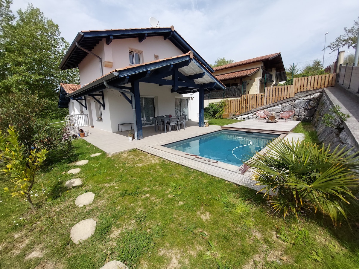 The exterior of a two-story house is displayed, featuring a wooden deck adjacent to a rectangular salt-water pool. Seating is provided for outdoor dining under a shaded area. Lush greenery surrounds the property, with stepping stones leading through the garden.