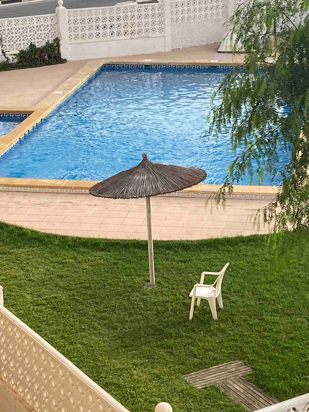 A swimming pool is surrounded by a well-maintained lawn. A grass area features a single white chair and a thatched umbrella providing shade, creating a serene spot for relaxation by the water.