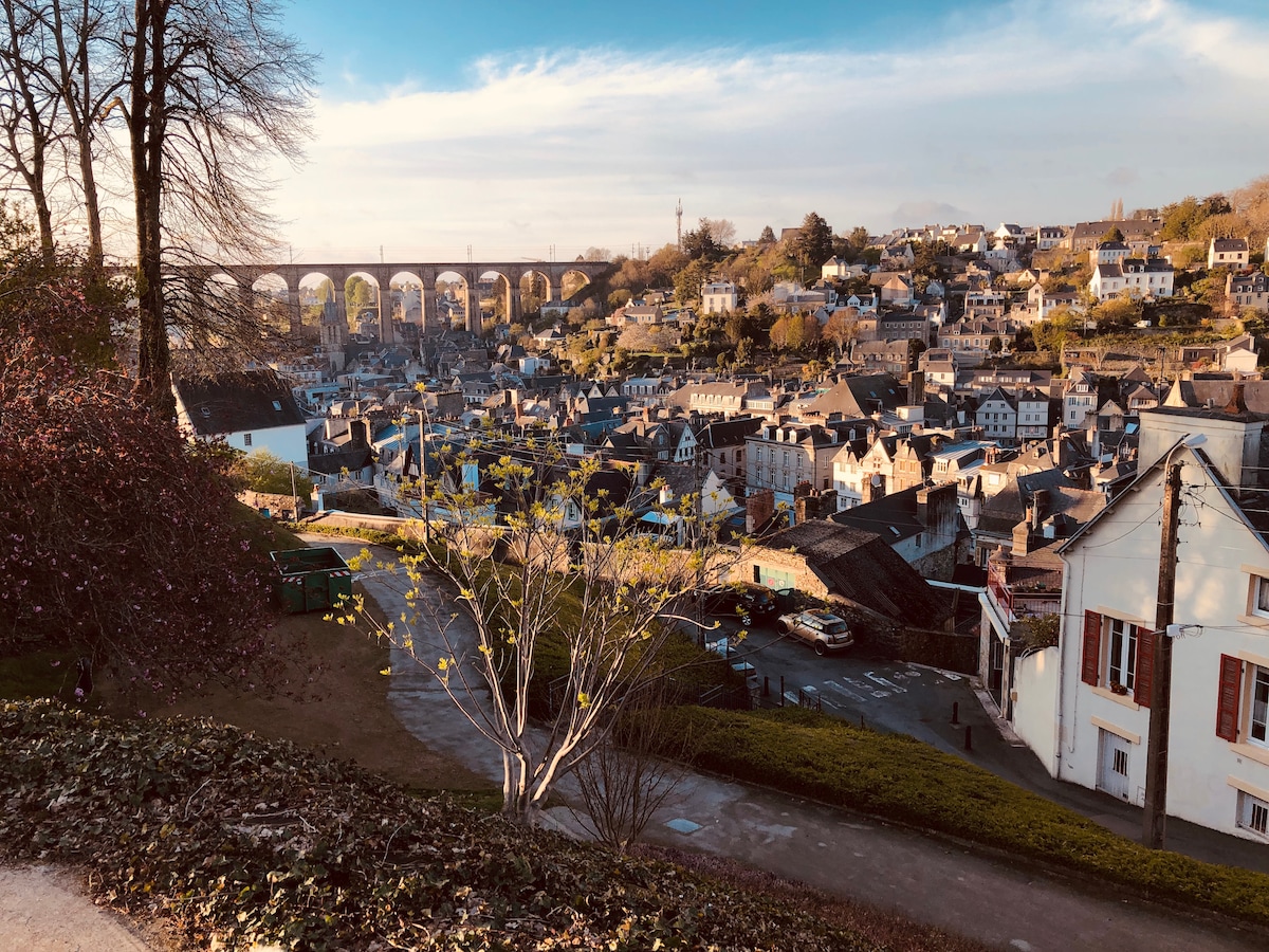 An expansive view of Morlaix showcases the town's charming rooftops and the prominent viaduct in the background. Lush trees and greenery are visible in the foreground, framing the scene, while the light blue sky adds a serene touch to the vibrant landscape.