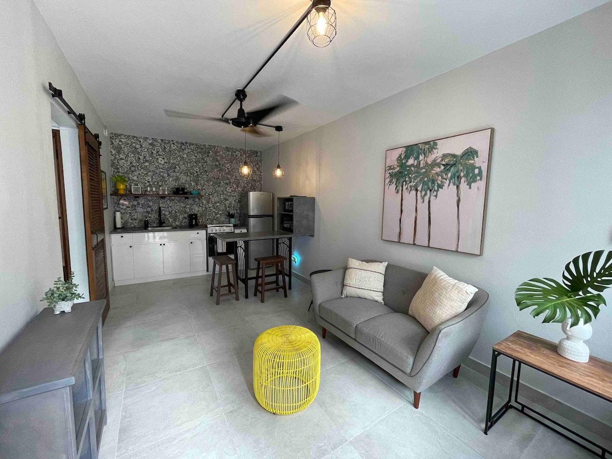 A modern living space features a light gray sofa with decorative pillows and a small yellow table. A kitchenette with stainless steel appliances and white cabinetry is visible in the background. Decor includes a tropical-themed wall art and a green plant in the corner.