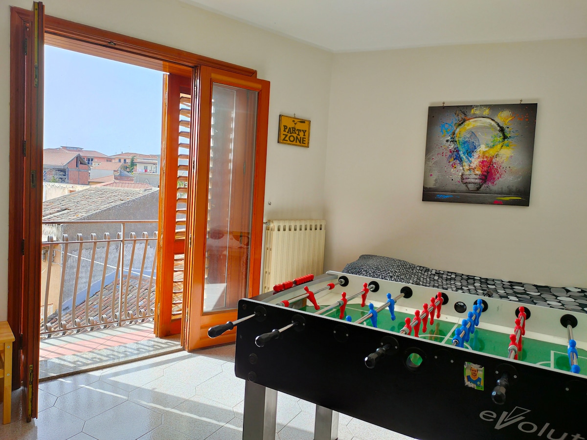 A light-filled room features a foosball table in the foreground and a spacious bed with patterned bedding. Large glass doors open to a balcony, providing a view of the surroundings. Natural light enhances the bright atmosphere, while a radiator is positioned on the wall.