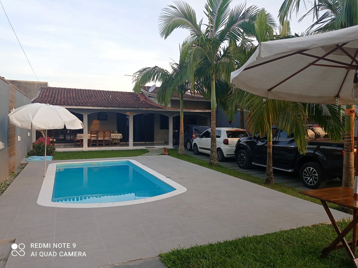 A private backyard area features a rectangular pool surrounded by tile flooring and lush green grass. Palm trees provide shade alongside a covered seating area with a large wooden table. Multiple vehicles are parked nearby, indicating convenient access.