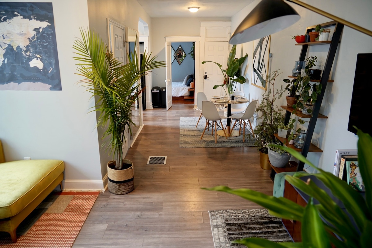 A bright living area features a green couch and a large plant, creating a vibrant atmosphere. A dining table surrounded by white chairs sits in the background, while various plants enhance the space. Art pieces and a patterned rug add color and texture.