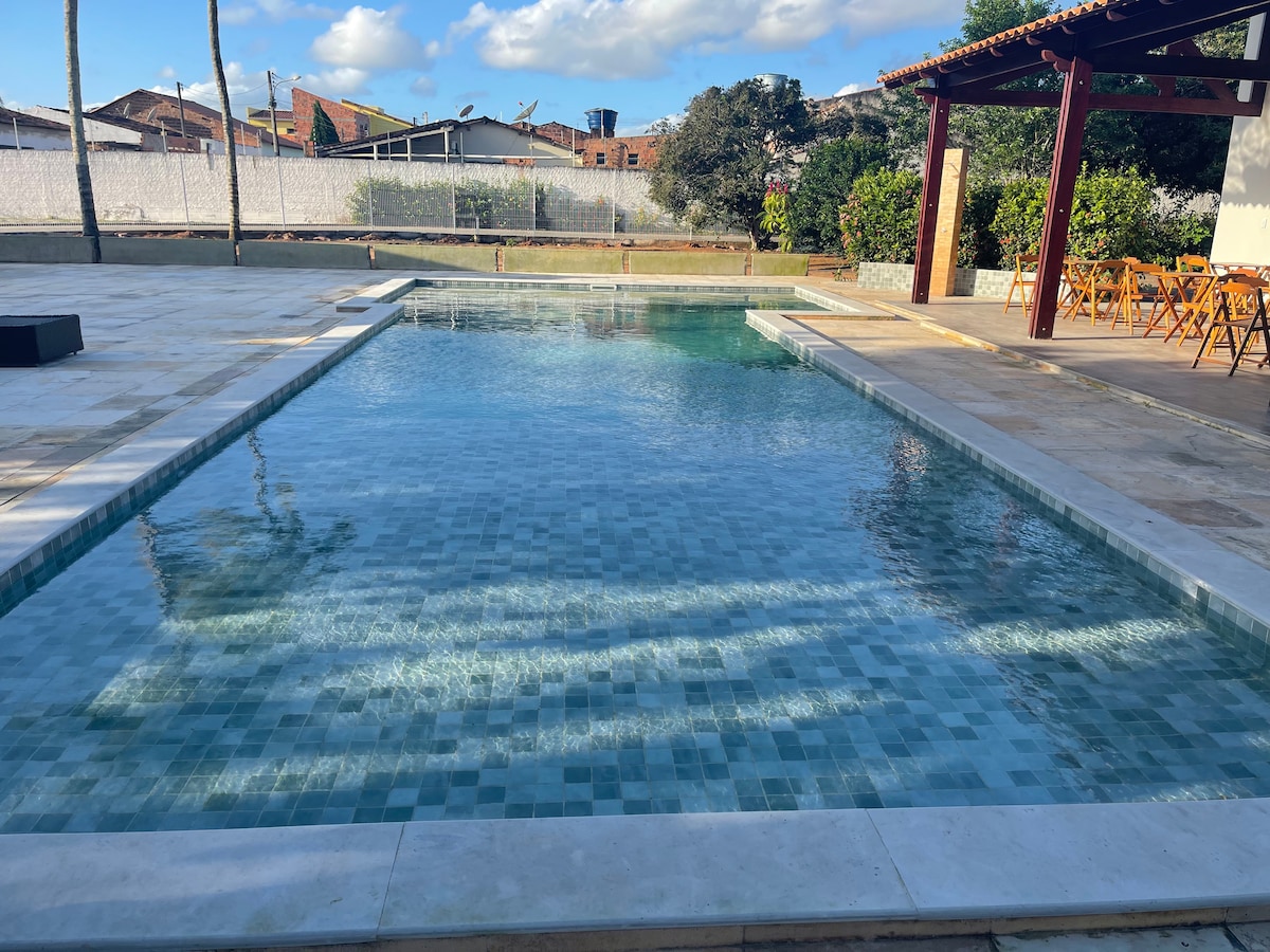 A clear swimming pool features tiled edges and is surrounded by open space. Sunlight reflects off the water's surface, showcasing various shades of blue. A shaded terrace with wooden furniture is visible in the background, alongside greenery and a fence.