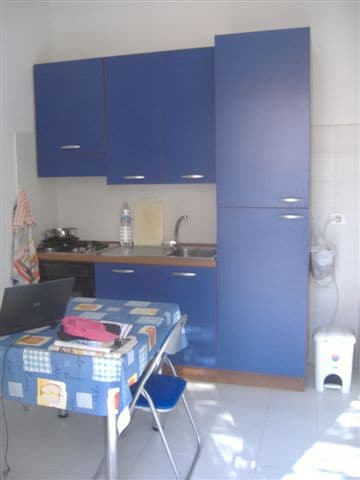 The kitchen area showcases blue cabinetry and a countertop with a sink and stove. A table is set up with chairs, and a laptop is placed on the table. A water bottle is visible, along with essential kitchen utilities in a bright, clean environment.