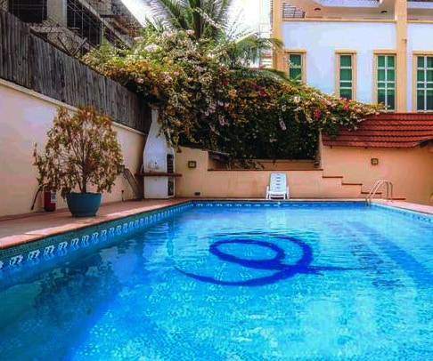 A clear swimming pool is framed by lush greenery and flowering plants, providing a serene atmosphere. A single white lounge chair is positioned beside the pool, while the surrounding area is tiled and well-maintained, creating a welcoming outdoor environment.