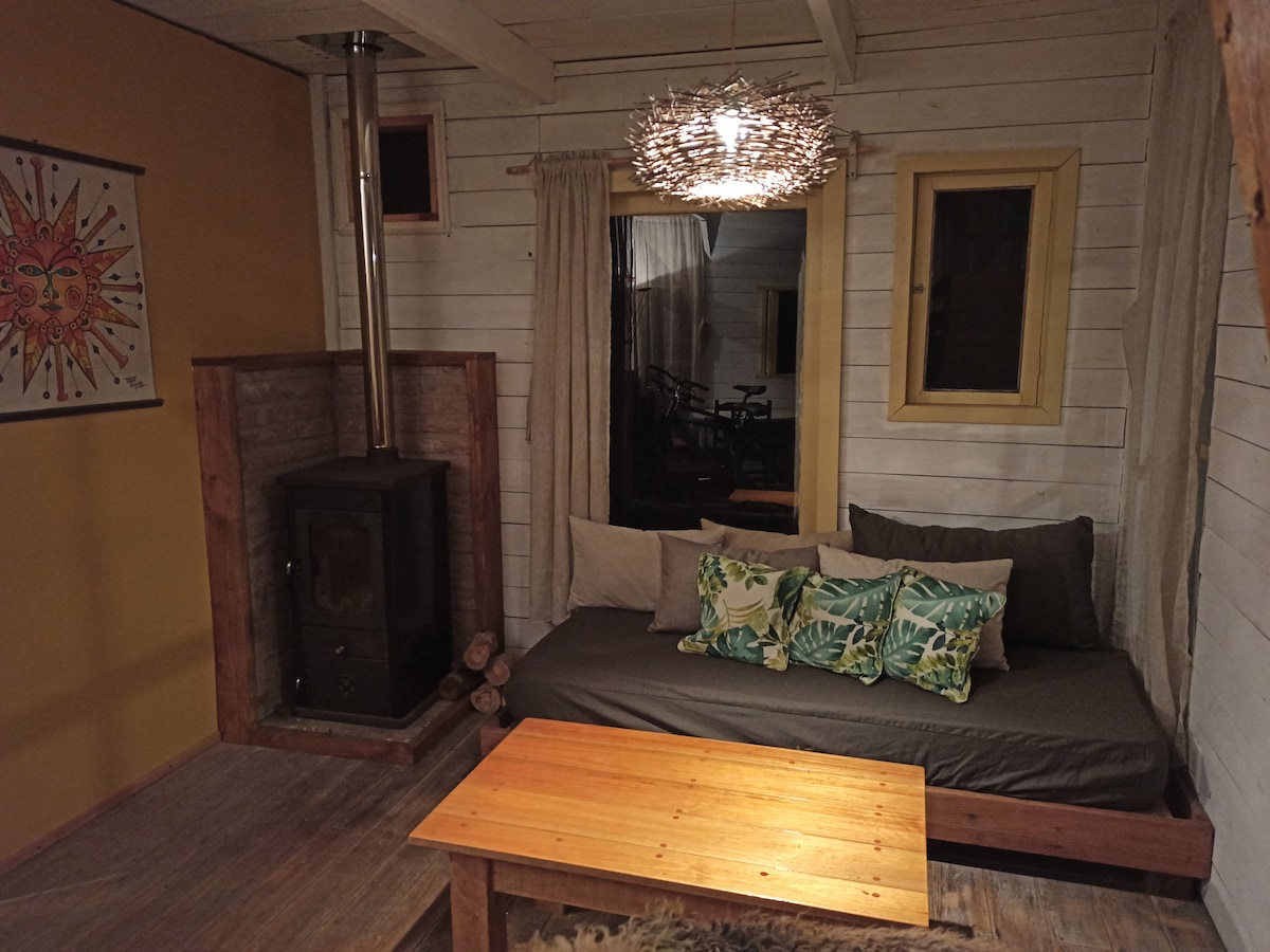 An inviting living area features a dark seating arrangement adorned with tropical decorative pillows. A wooden coffee table rests on the floor, while a metal stove stands adjacent to it. Natural light enters through a window, and a unique light fixture hangs overhead.