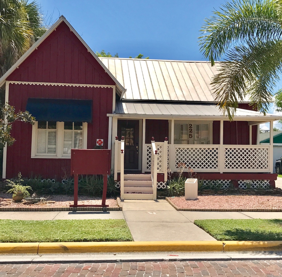 A charming red cottage with white accents and a metal roof is showcased. The front porch features decorative railings with seating options, while tropical plants are positioned alongside the walkway. A clear blue sky is visible overhead, emphasizing the inviting exterior.