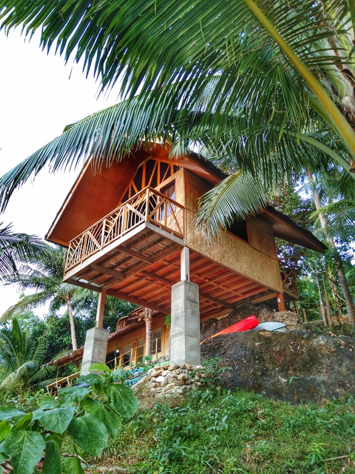 A wooden structure is elevated on stilts, surrounded by lush palm trees. Its front features a balcony with wooden railing, providing a glimpse of the natural environment. The earthy tones of the building complement the greenery, and a kayak rests nearby on the ground.
