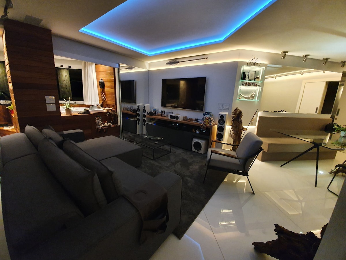 A modern living space features a sectional sofa complemented by sleek chairs and a glass dining table. Soft blue LED lighting accents the ceiling, enhancing the contemporary design. Large screens are mounted on the walls, with decorative elements integrated into the room for visual interest.