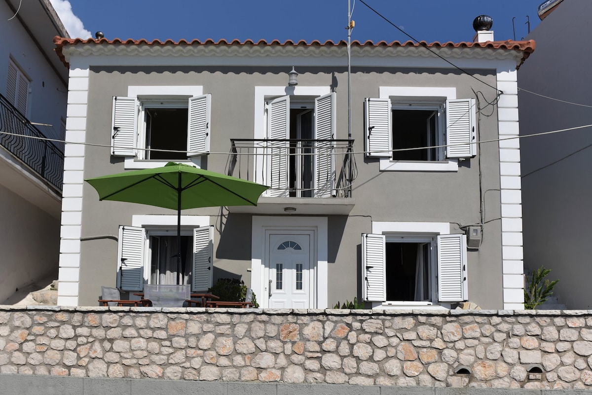 The exterior of the stone house features white shutters and an inviting front door. A green umbrella is positioned over outdoor seating, which is visible on a stone terrace. Above, a balcony with railings adds charm to the two-story structure.