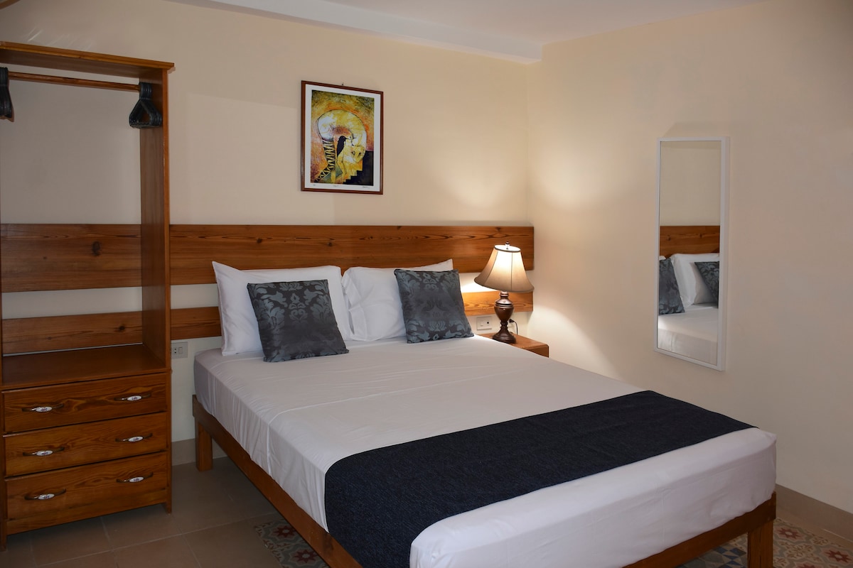 A well-lit bedroom is furnished with a spacious bed, soft white linens, and dark decorative pillows. A nightstand with a lamp stands next to the bed. A wooden wardrobe and dresser provide ample storage, while an art piece and a mirror are displayed on the walls.