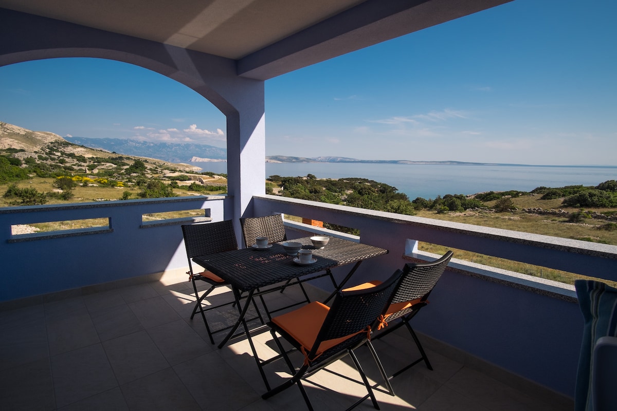 A spacious balcony offers a coastal view, featuring a table with two chairs and additional seating. Sunlight illuminates the area, with a serene backdrop of blue skies and distant hills along the shoreline. The balcony is framed by an arched entrance, enhancing the inviting space.