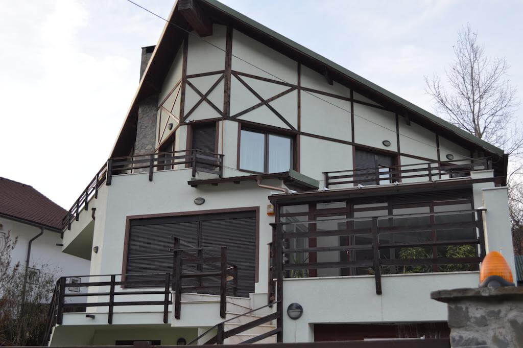 The exterior of a multi-level villa features a combination of wooden and stone accents. Large, modern windows are framed by a balcony that extends across the front side. A well-maintained pathway leads to the main entrance, complemented by lush greenery at the base.