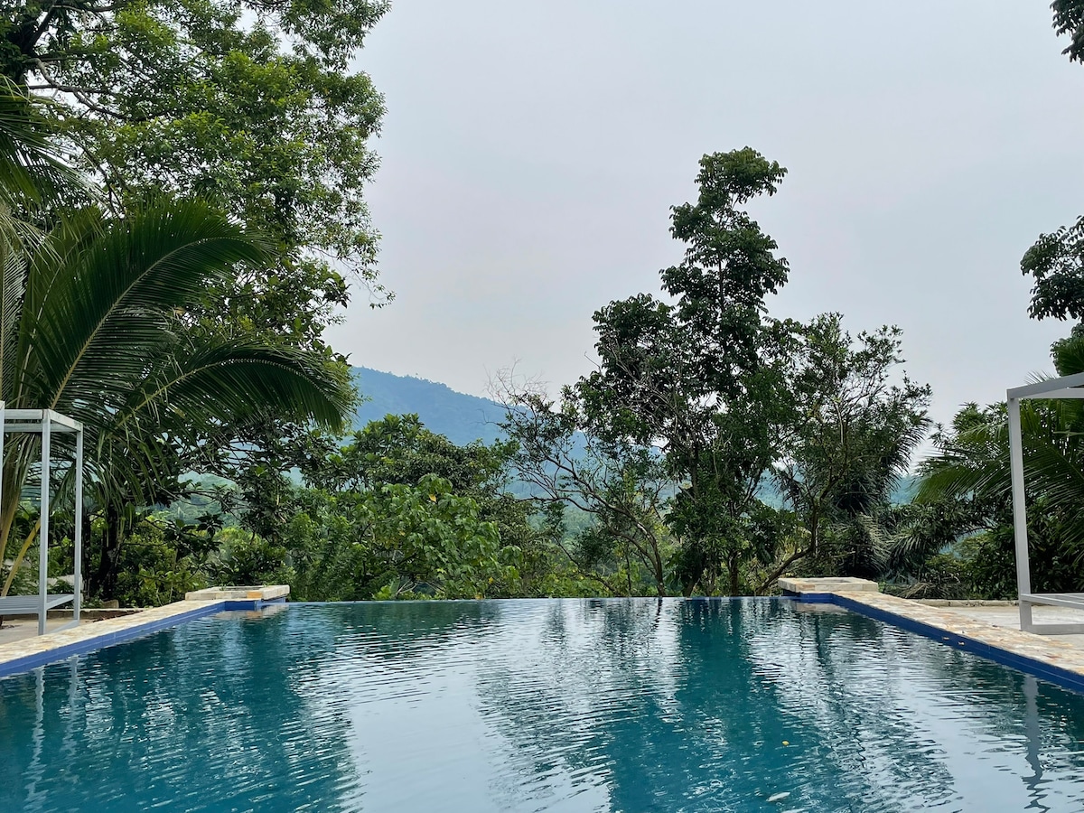 An infinity pool extends into the lush greenery, framed by tropical foliage. The tranquil water reflects the overcast sky, while scenic hills are visible in the background, enhancing the sense of seclusion and natural beauty.