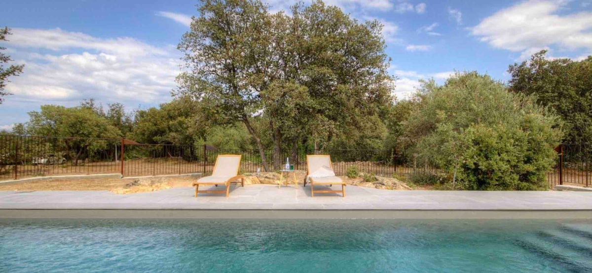 Two lounge chairs are positioned beside a tranquil swimming pool, surrounded by lush greenery. A large tree offers shade in the background, while a fence delineates the outdoor area, providing a sense of privacy and nature.
