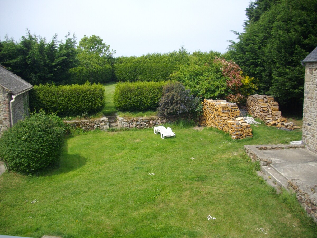 An expansive garden is showcased, featuring well-maintained grass and bordered by neatly trimmed hedges. A single lounge chair is situated on the lawn, and a stack of firewood lies against the stone wall, contributing to the serene outdoor setting.