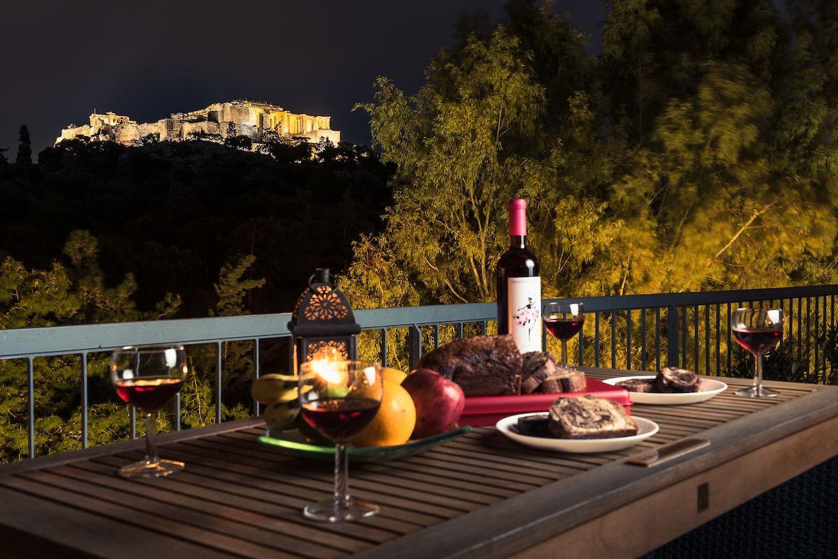 An outdoor dining table is set with red wine glasses and a bottle of wine. A platter of fruits and a dessert rests on the table, with the illuminated Acropolis in the background, surrounded by lush greenery.