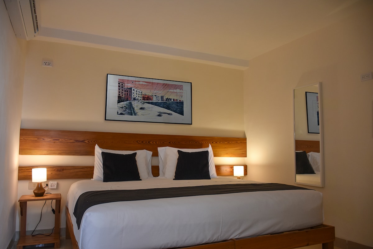 A spacious sleeping area features a large bed adorned with crisp white linens and dark accent pillows. Soft bedside lighting complements the modern artwork hanging above the headboard, while a full-length mirror reflects the serene ambiance of the room.