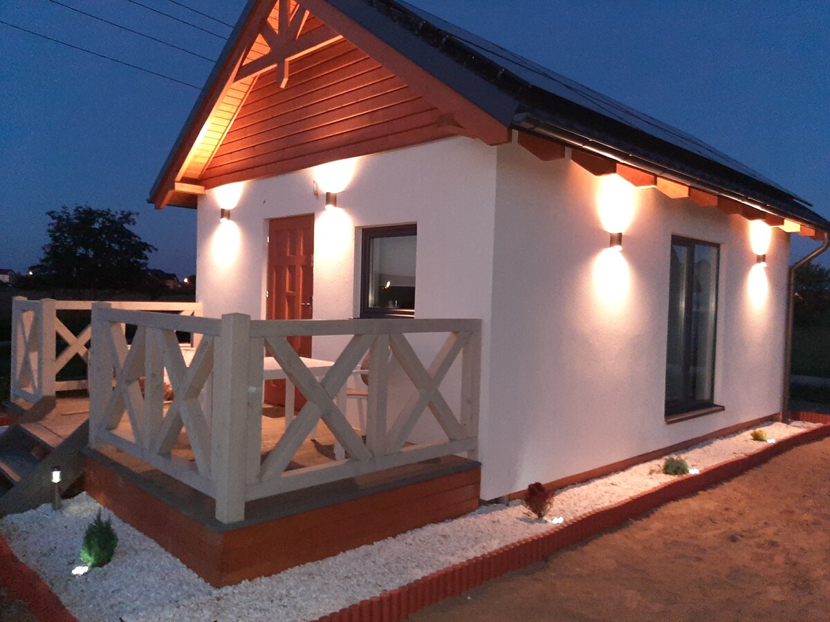 The exterior of a small, modern cottage is showcased at dusk, featuring soft lighting that highlights the architectural details. A spacious front porch is framed by a decorative railing, while a large window allows glimpses of the interior. Lush landscaping accents the pathway.