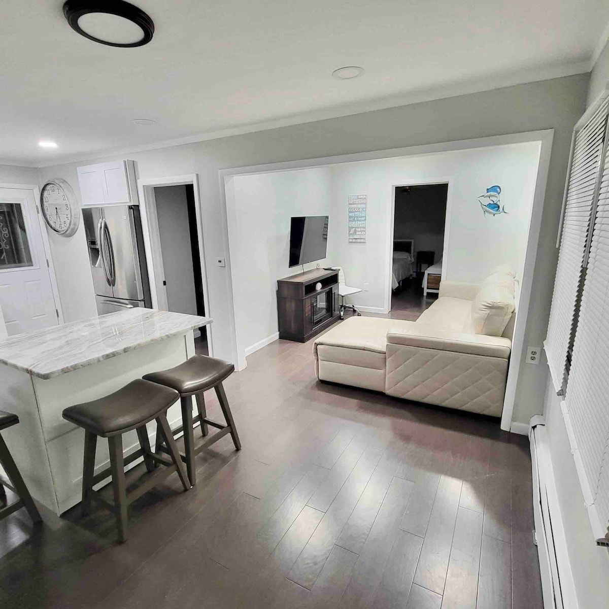 A modern living space features an open layout with a cozy sofa positioned near a television stand. A marble kitchen island with bar stools enhances the area, while soft lighting and light-colored walls contribute to a bright atmosphere. Doorways lead to additional spaces.