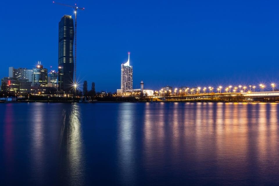 A nighttime view captures the skyline of Vienna, with illuminated buildings reflecting off the calm waters of the Danube River. The arrangement of lights creates a serene atmosphere, contrasting with the dark blue sky above.
