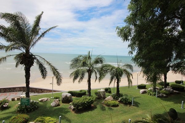 Lush greenery and palm trees frame a view of the sandy beach and the Gulf of Thailand. The gentle waves can be seen in the distance, while small rocks and manicured shrubs adorn the shoreline. A calm sky reflects the serene coastal atmosphere.