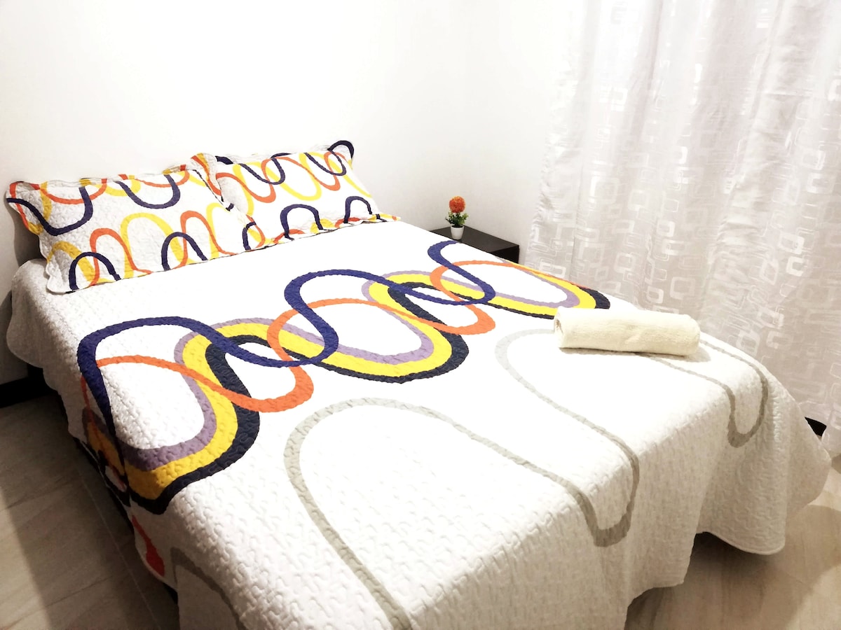 A cozy sleeping area features a double bed adorned with a colorful quilt and matching pillows. Natural light filters through a sheer white curtain, creating a bright atmosphere. A small plant sits on a bedside table, adding a touch of greenery to the space.