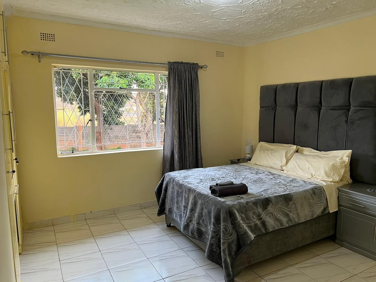 A well-lit sleeping area features a spacious bed with neutral bedding and a decorative grey throw. Natural light enters through a large window, framed by dark curtains, while a small bedside table sits beside the bed.