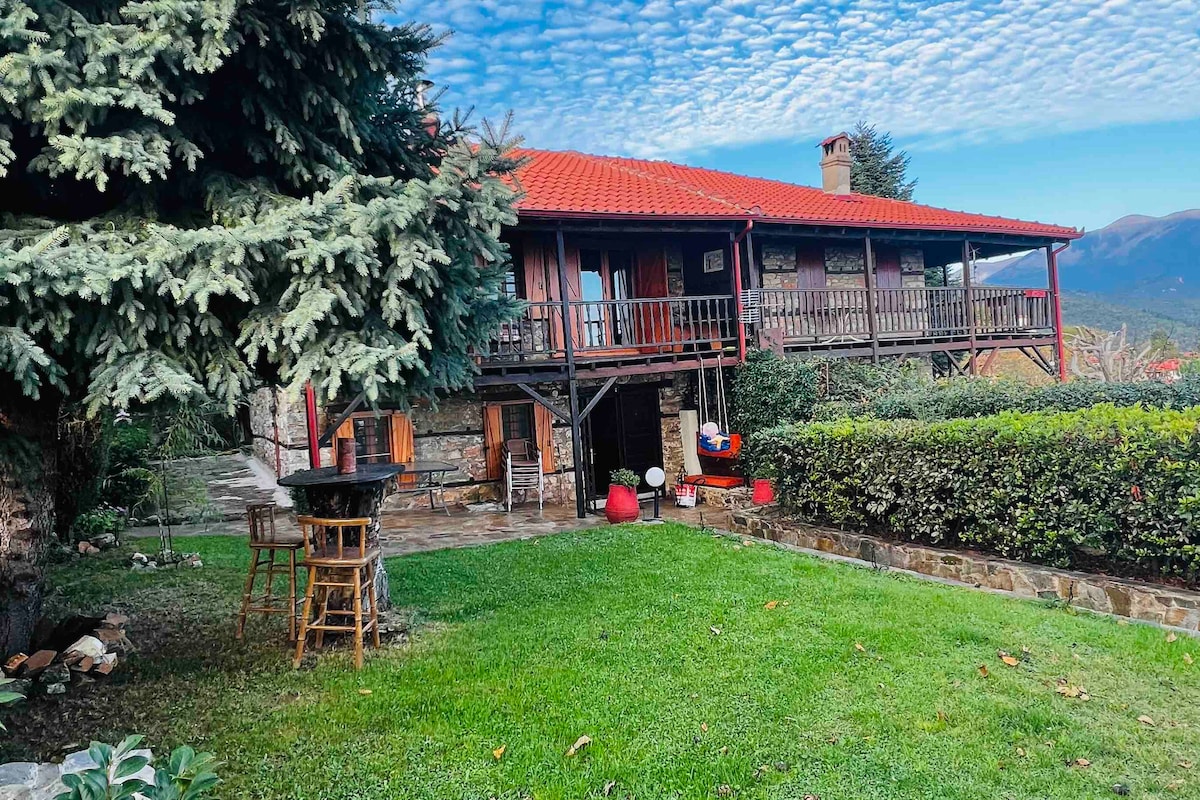 A charming villa is surrounded by lush greenery and features a red-tiled roof. A spacious porch offers seating and views of the garden, while stone pathways lead through the well-maintained landscape. The nearby trees provide shade and enhance the serene atmosphere.