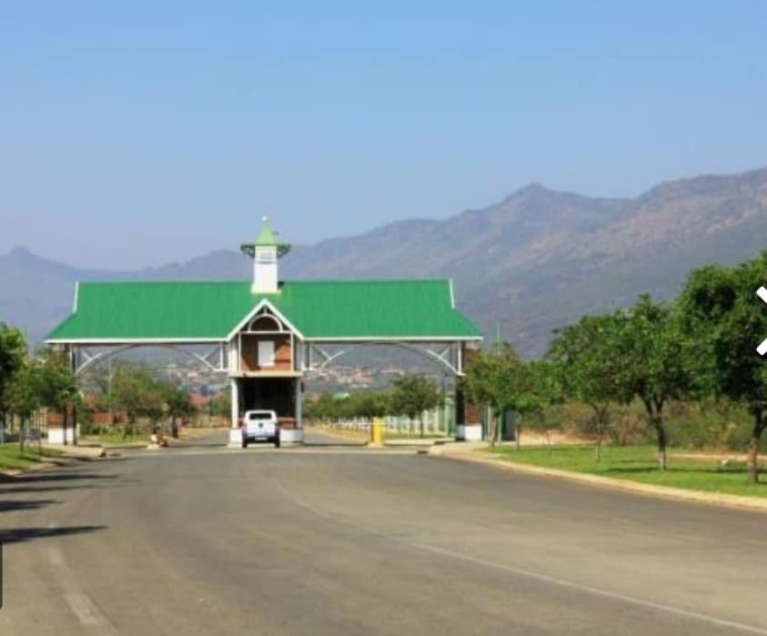 An entrance gate to a secure estate is prominently featured, characterized by a peaked green roof and a white structure. Tree-lined pathways lead to the gate, with distant mountains visible under a clear blue sky.