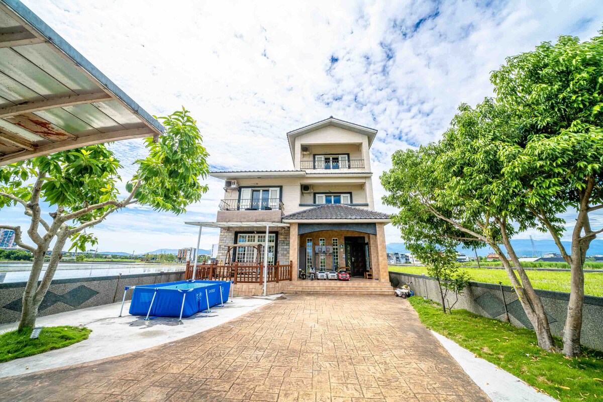 A three-level house is set against a scenic backdrop, showcasing a spacious driveway lined with greenery. An outdoor area features a blue pool surrounded by concrete and grass, with large windows offering views of the landscape beyond.