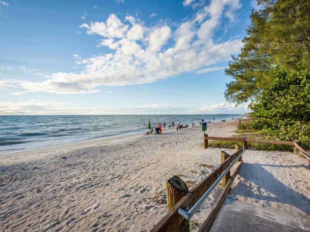 The image showcases a sandy beach with gentle waves lapping at the shore. Lush greenery borders the beach, and a pathway leads toward the water's edge. A few beachgoers are seen, enjoying the serene surrounding and expansive sky scattered with clouds.